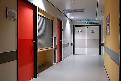 Couloir d'hôpital avec portes rouges et doubles portes blanches au fond.