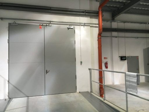 Porte grise en métal située à l'intérieur d'un bâtiment industriel. Mur blanc et tuyauterie rouge.