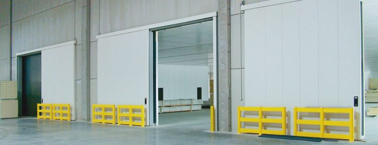 Des portes coulissantes blanches ouvertes dans un entrepôt avec des barrières jaunes.