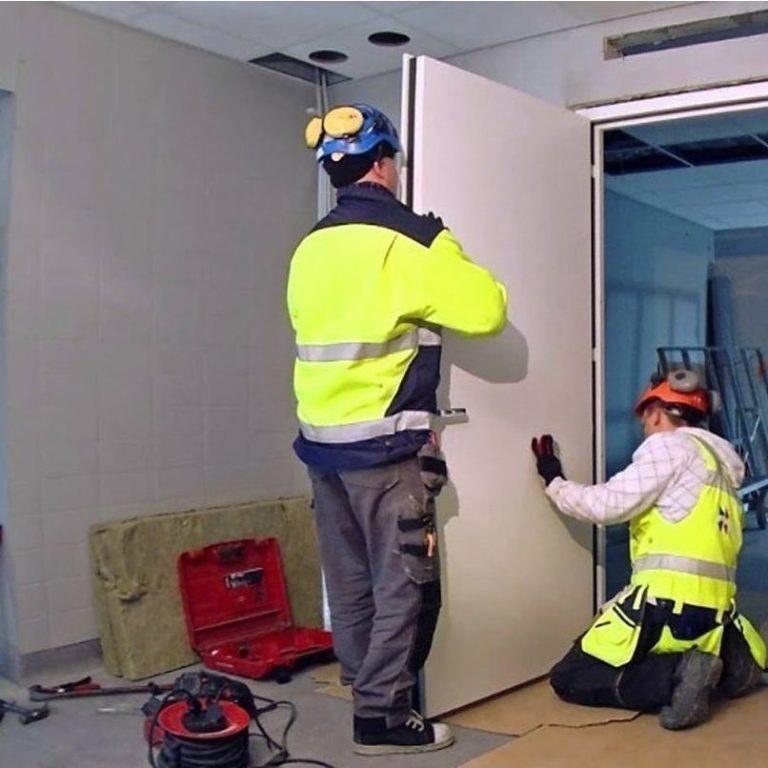 Deux travailleurs en tenue de sécurité installent une porte dans un bâtiment.