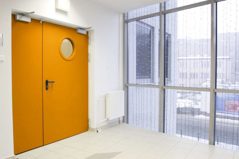 Porte orange avec un hublot dans un couloir moderne et lumineux. Fenêtres à côté.