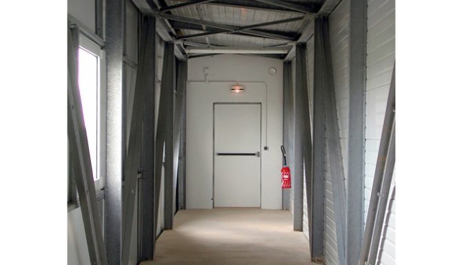 Couloir étroit avec murs en métal et une porte fermée au fond.