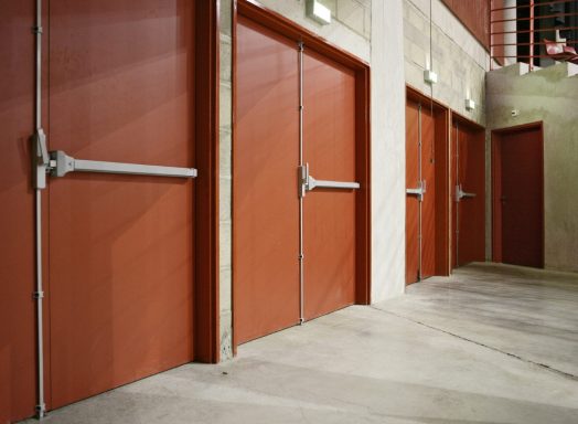 Quatre portes de secours rouges avec poignées en métal, sur un sol en béton.